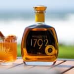 A glass of cocktail with a cinnamon stick and orange slice beside a bottle of 1792 bourbon on an outdoor table.