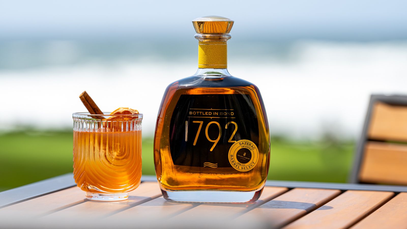 A glass of cocktail with a cinnamon stick and orange slice beside a bottle of 1792 bourbon on an outdoor table.
