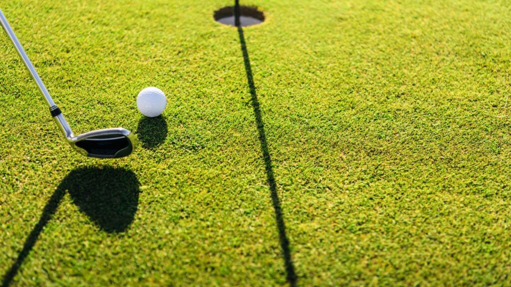 A golf club prepares to putt a golf ball towards a hole on a green, with shadows cast by the club and flagstick.