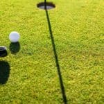 A golf club prepares to putt a golf ball towards a hole on a green, with shadows cast by the club and flagstick.