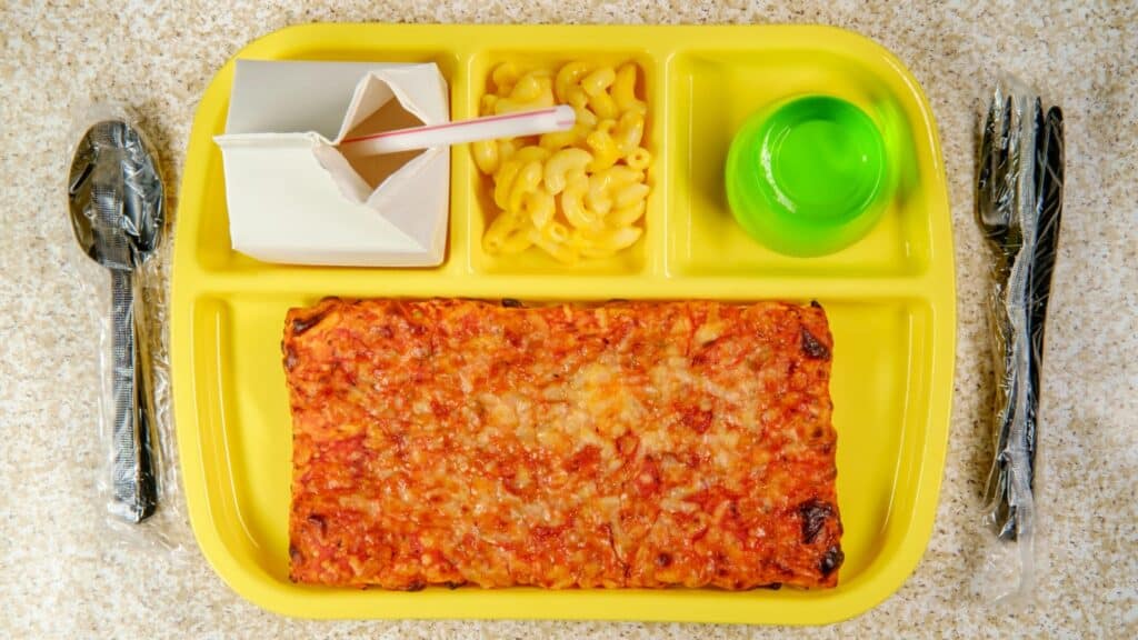 Yellow lunch tray with rectangle pizza, macaroni and cheese, green gelatin dessert, milk carton with straw, and wrapped plastic fork and spoon on a granite surface.