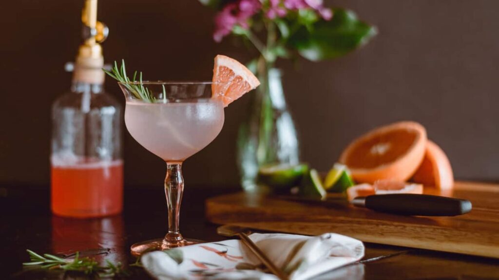 A cocktail garnished with a grapefruit slice and rosemary sits on a table with a napkin, a sliced grapefruit, lime wedges, a knife, and flowers in the background&mdash;evoking nostalgic vibes reminiscent of colorful 80s and 90s school cafeteria foods.