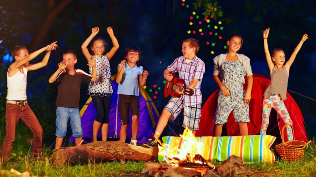 Seven children stand and dance by a campfire with tents and string lights in the background; one child plays a guitar, creating the kind of summer camp traditions campers remember for years to come.