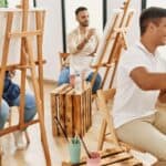 A group of adults sit at easels in a bright room, painting on canvases. Wooden stools and art supplies are visible.