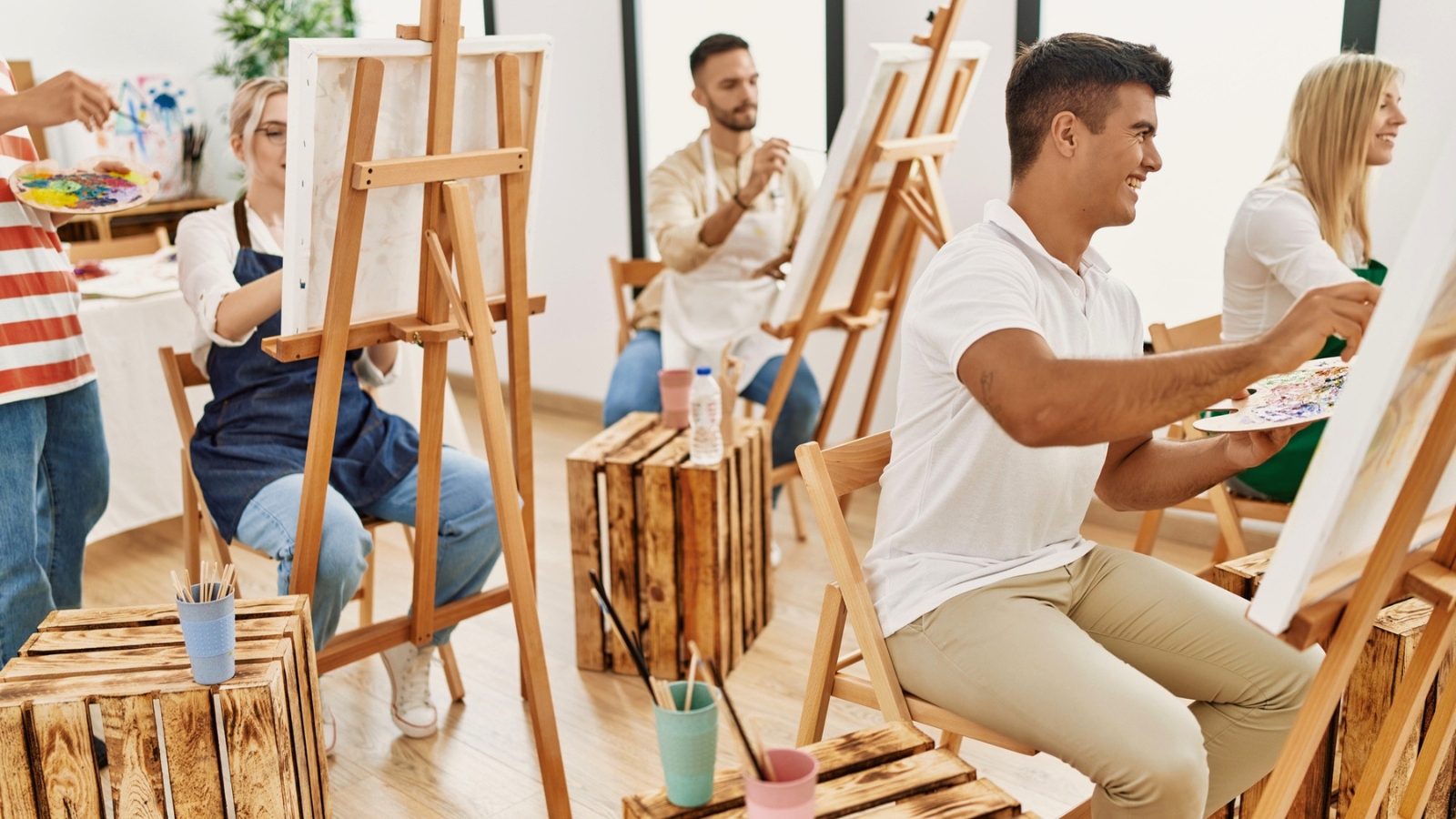 A group of adults sit at easels in a bright room, painting on canvases. Wooden stools and art supplies are visible.
