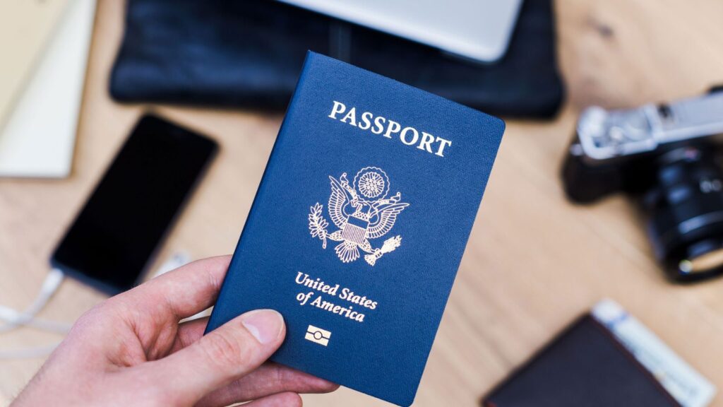 A hand holds a United States passport, perhaps anticipating upcoming adventures despite current passport wait times. A smartphone, camera, and other travel items are on a wooden table in the background.