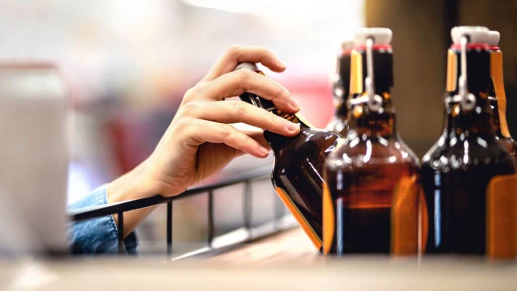 A person&rsquo;s hand is reaching for a brown glass bottle from a row of similar bottles on a store shelf, carefully considering options while being mindful of state alcohol laws.