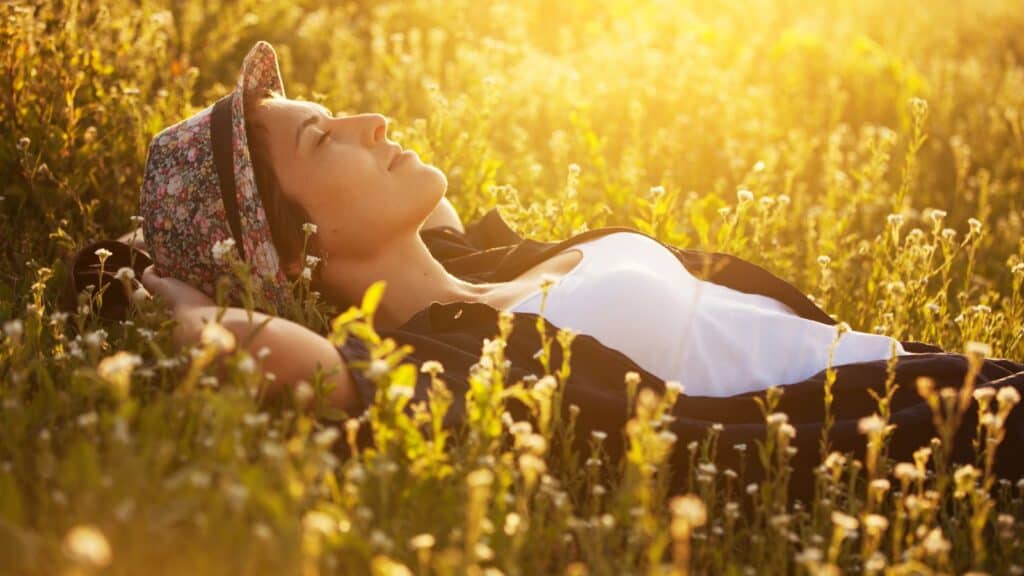 Person wearing a floral hat and white shirt lies on their back in a sunlit field of wildflowers, eyes closed, enjoying the warm light.