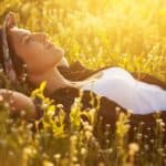 Person wearing a floral hat and white shirt lies on their back in a sunlit field of wildflowers, eyes closed, enjoying the warm light.