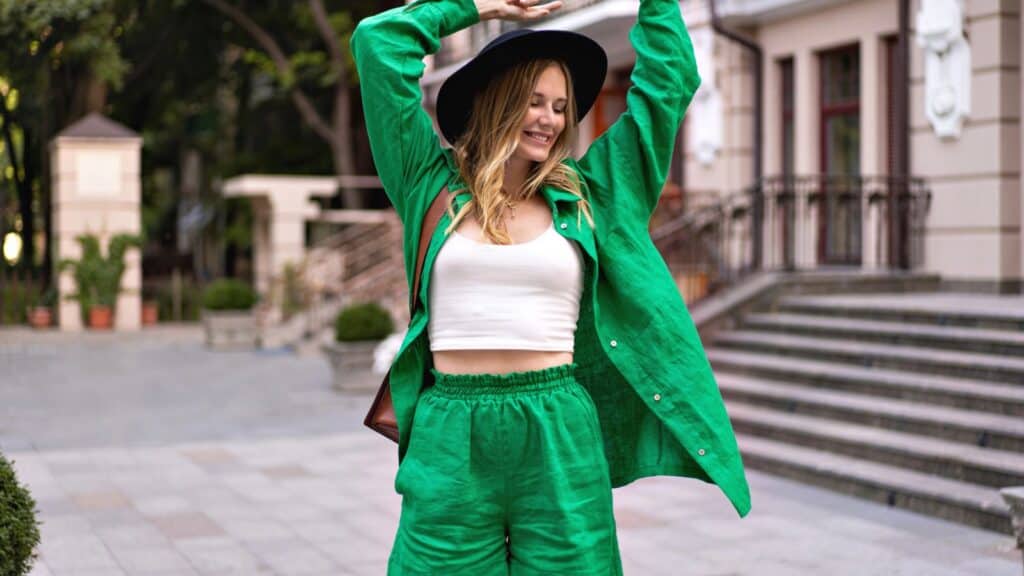 Person wearing a green outfit and black hat stands outdoors with arms raised, smiling, near steps and a building.