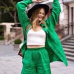 Person wearing a green outfit and black hat stands outdoors with arms raised, smiling, near steps and a building.