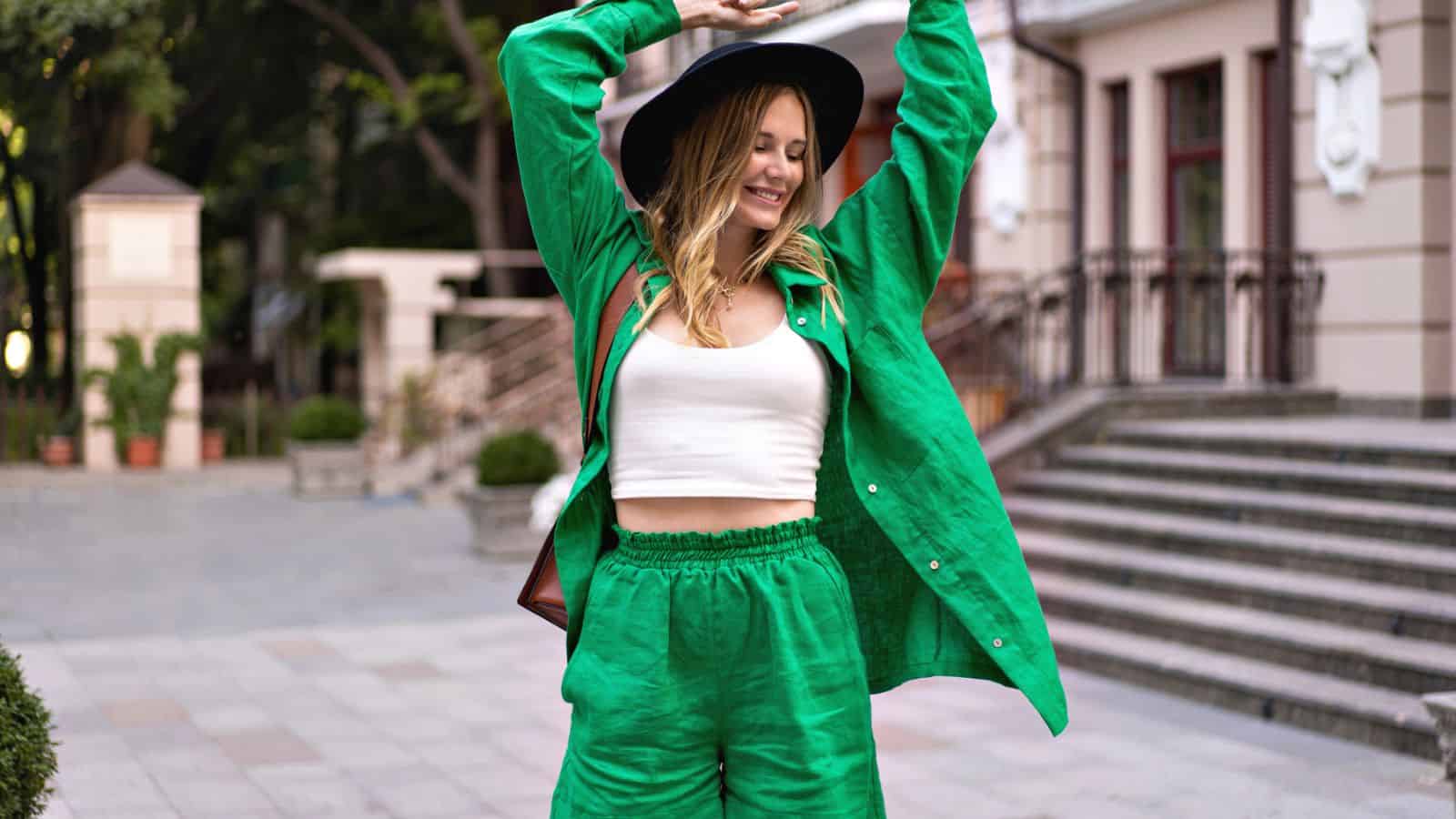 Person wearing a green outfit and black hat stands outdoors with arms raised, smiling, near steps and a building.