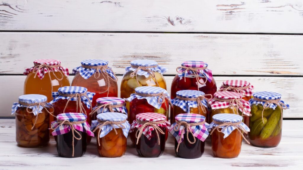 Fifteen jars of assorted homemade preserves&mdash;foods US customs may confiscate&mdash;with checkered fabric lids and twine, arranged in rows on a wooden surface against a white wood background.