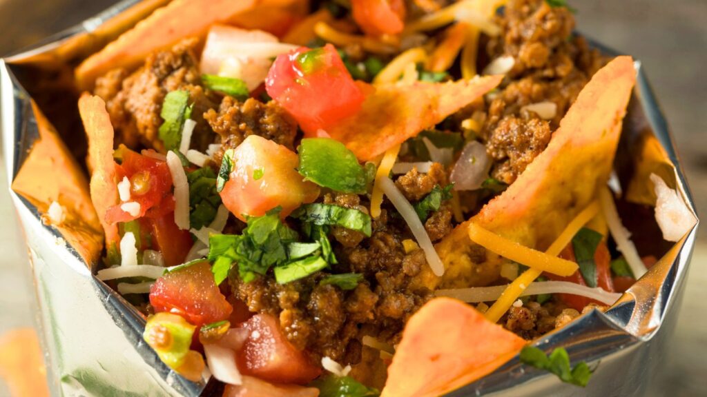 Close-up of a bag of chips filled with seasoned ground beef, shredded cheese, diced tomatoes, onions, and chopped cilantro.