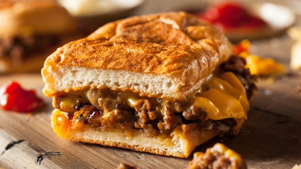 A close-up of a toasted sandwich filled with melted cheese and ground beef, with a bite taken out of it, on a wooden surface.