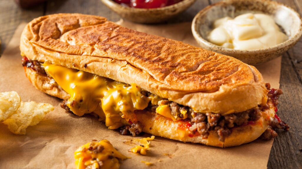 A toasted cheesesteak sandwich filled with melted cheese and chopped beef sits on brown paper, with potato chips and dipping sauces in the background.