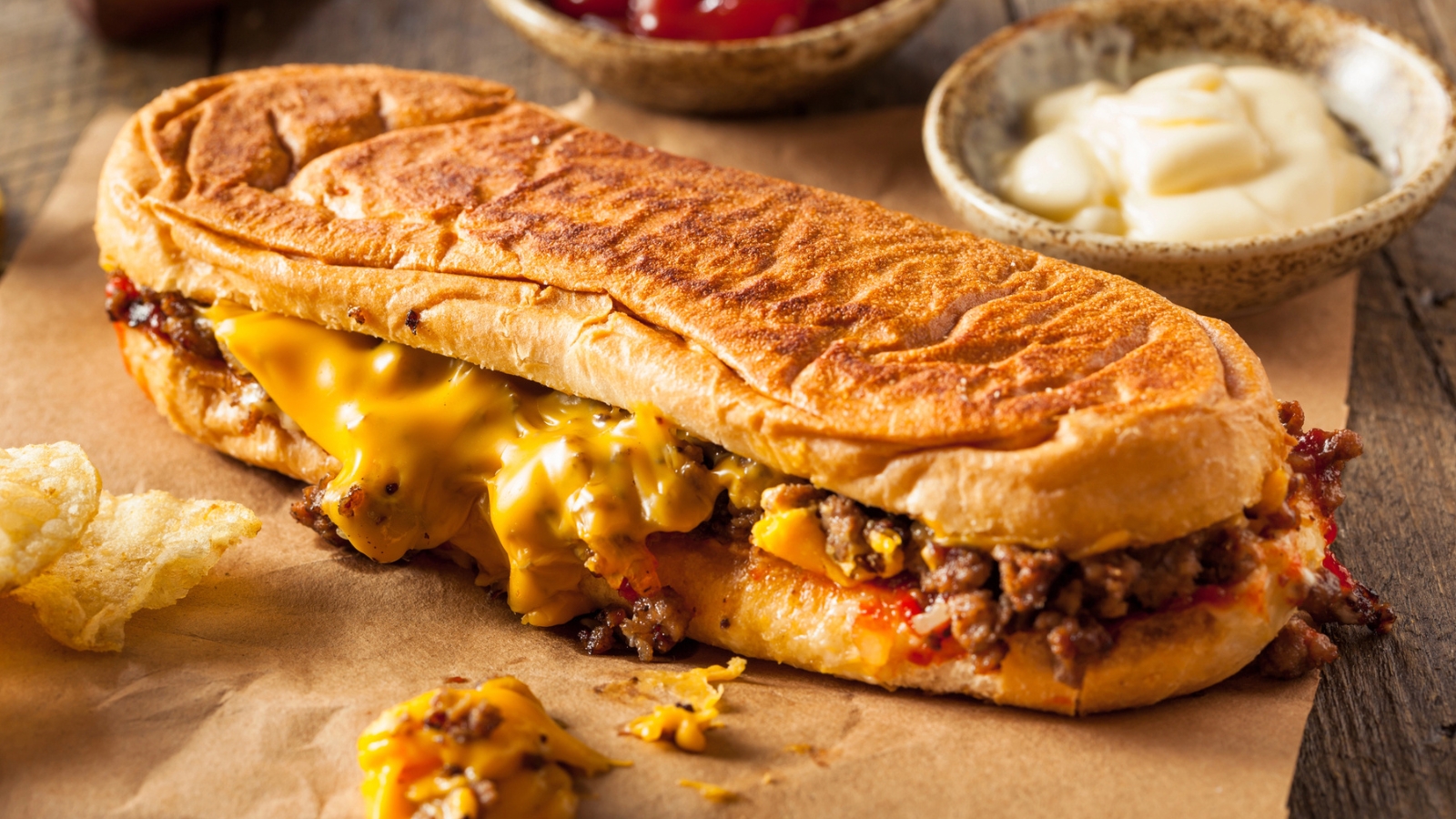 A toasted cheesesteak sandwich filled with melted cheese and chopped beef sits on brown paper, with potato chips and dipping sauces in the background.