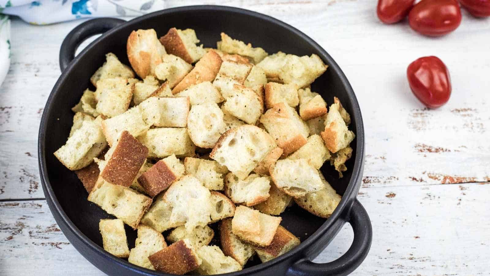 Black bowl filled with golden-brown croutons on a white wooden surface, with a few cherry tomatoes nearby.