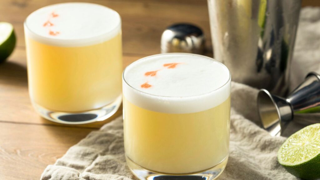 Two pisco sour cocktails with frothy tops and bitters, next to a cocktail shaker, jigger, linen cloth, and sliced lime on a wooden surface.