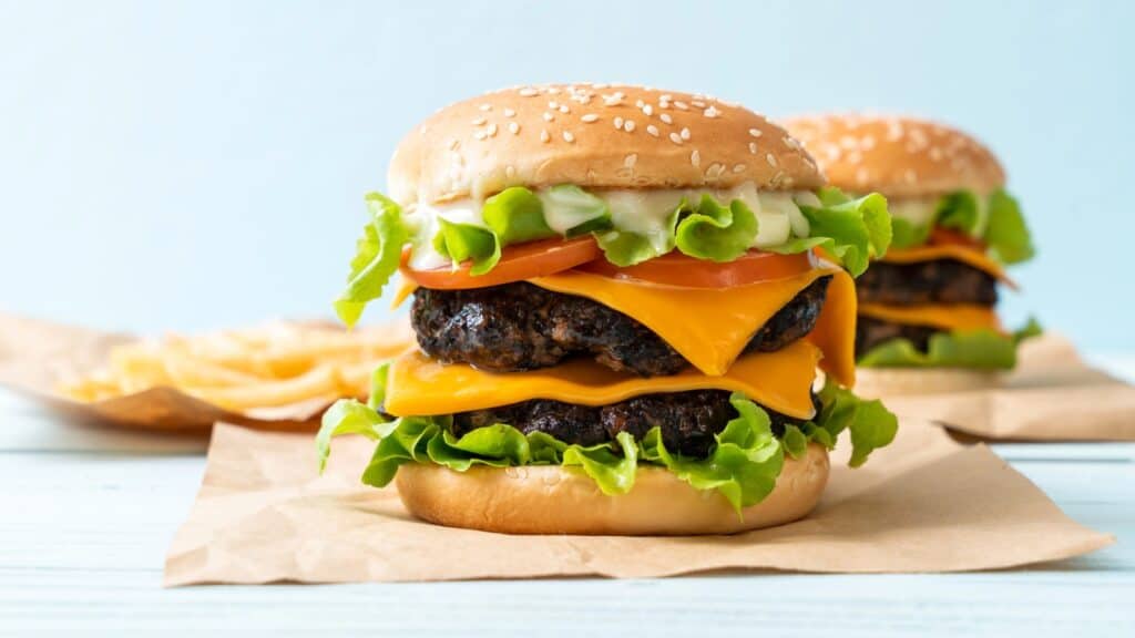A double cheeseburger with lettuce, tomato, and mayonnaise on a sesame seed bun sits on brown paper, with fries in the background.