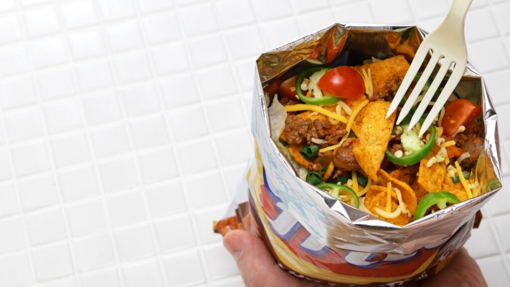 An open bag of corn chips filled with ground beef, shredded cheese, jalapeños, tomatoes, and lettuce; a plastic fork is inserted, and the background is white tile.