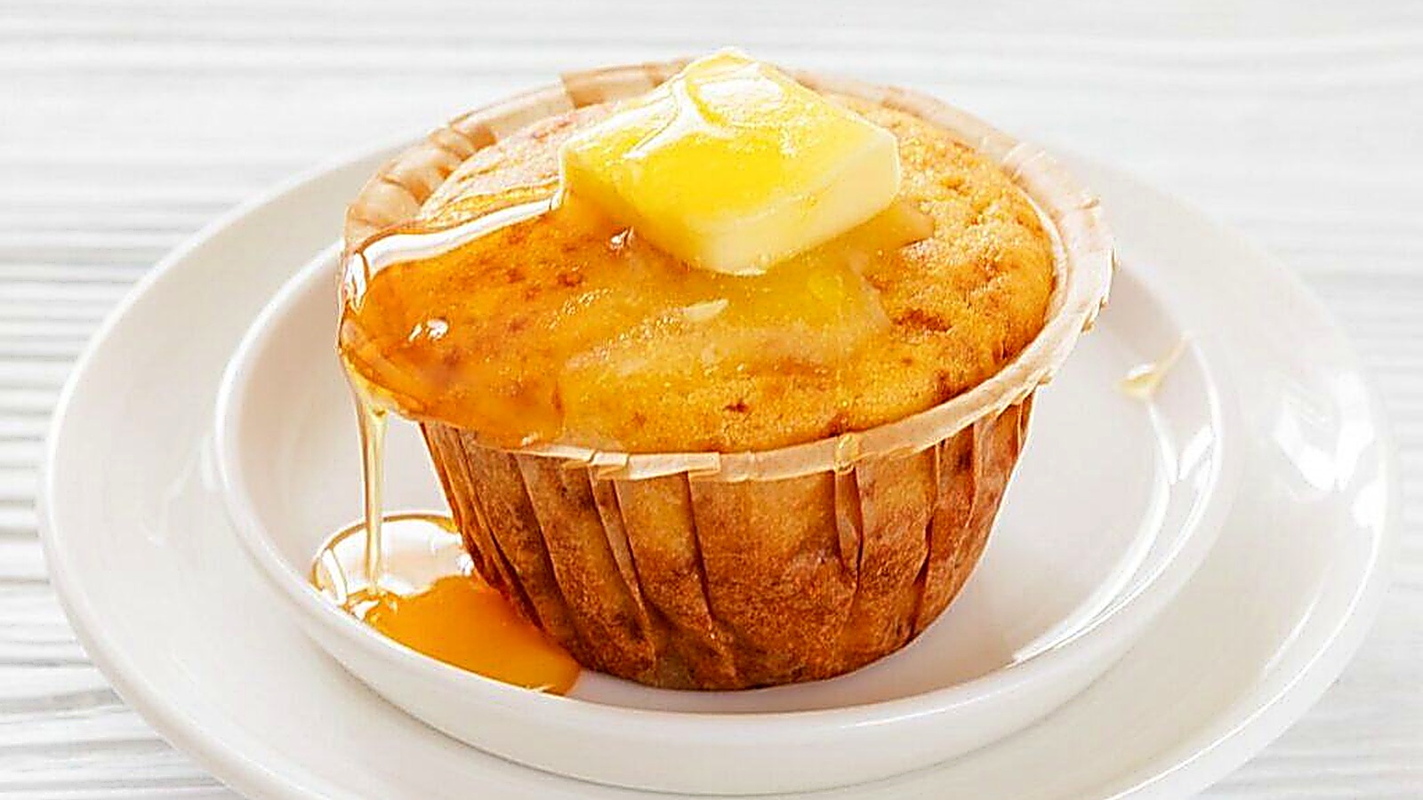 A Honey Cornbread muffin on a white plate, topped with a pat of butter and drizzled with syrup.
