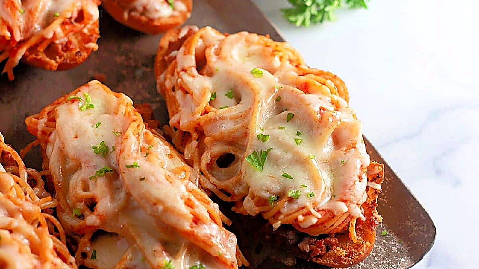 Toasted bread slices topped with spaghetti, meat sauce, melted cheese, and garnished with chopped parsley on a metal baking sheet.