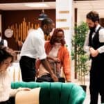 Hotel lobby scene with a guest being assisted by attentive hotel concierges near a green sofa, while another person reads documents and a receptionist stands at a desk in the background, ready to help with any request guests wish to ask.