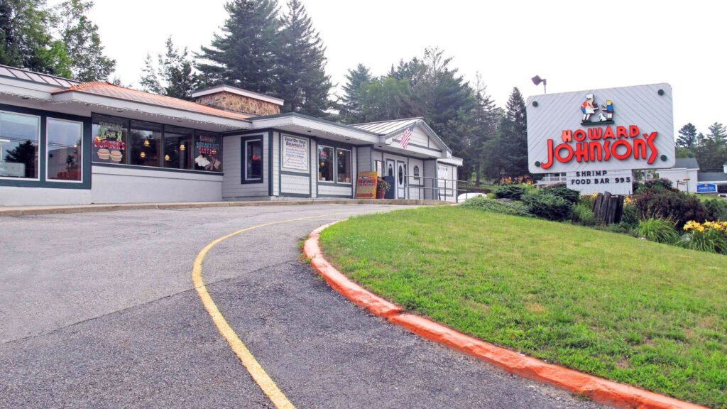 Exterior view of a Howard Johnson&rsquo;s restaurant&mdash;one of the iconic restaurant chains that faded away&mdash;with a large roadside sign and a curved driveway leading up to the entrance, surrounded by grass and trees in the background.