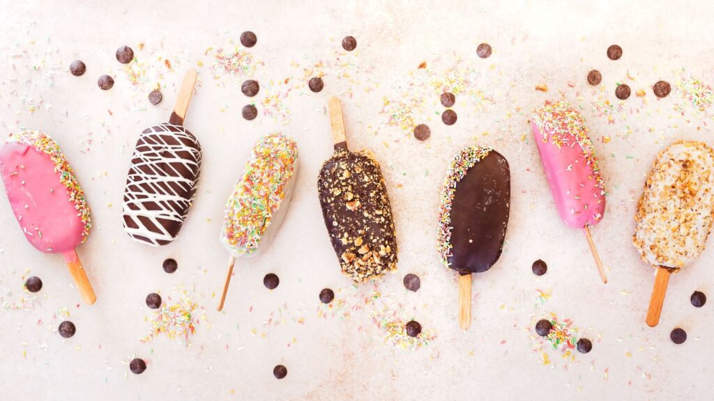 Seven decorated ice cream bars on sticks are arranged in a row, reminiscent of classic ice cream truck treats, with colorful sprinkles, chocolate chips, and crumbs scattered on a light background.