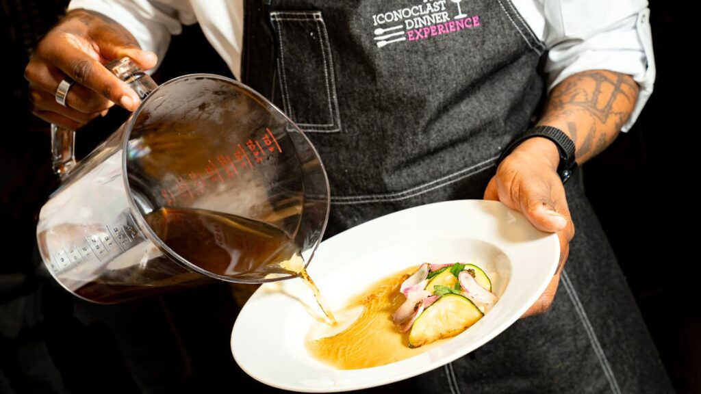 A person in a black apron pours broth from a measuring cup onto a plated dish of fish and vegetables.
