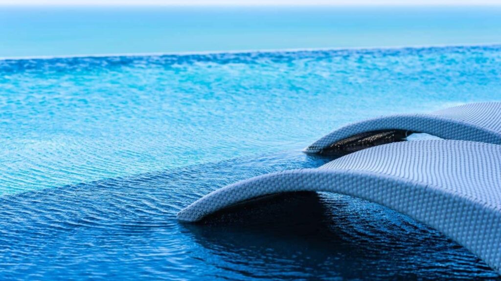 Two white lounge chairs partially submerged in a clear blue infinity pool overlooking the ocean on a sunny day.