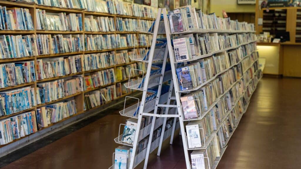 Rows of shelves filled with books and magazines are seen in a library or bookstore with a wooden counter in the background.
