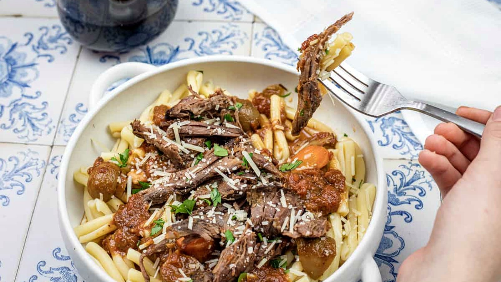 A hand lifting a fork with a bite of Italian Pot Roast Pasta on a bowl.