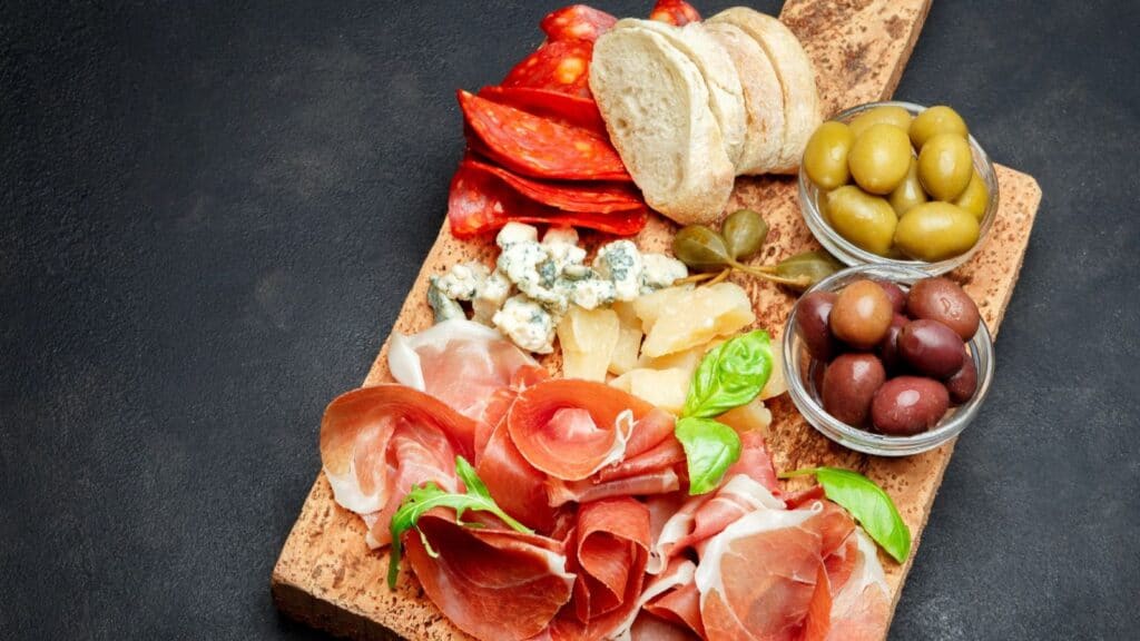 Charcuterie board with prosciutto, salami, cheese, bread slices, green and black olives, and fresh basil on a wooden board against a dark background.