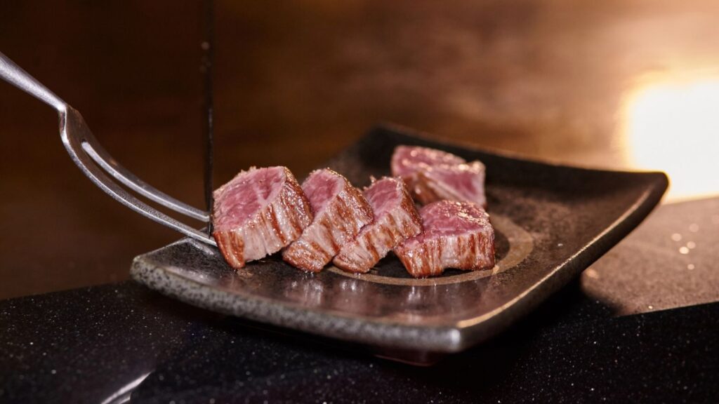 Five pieces of medium-rare steak are arranged on a dark ceramic plate, with a fork lifting one piece. The background is out of focus, subtly reminding us that sometimes foods are not what their name suggests&mdash;yet these perfectly cooked steaks look every bit as delicious as they sound.