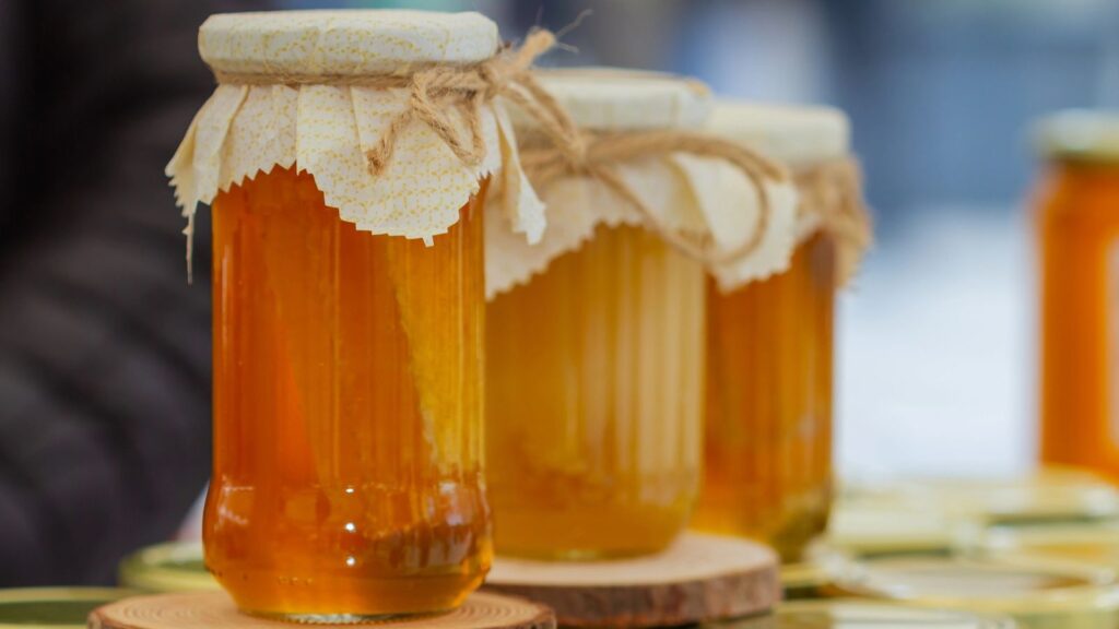 Three jars of honey with cloth and twine covers are displayed on a wooden surface, with a blurred background.