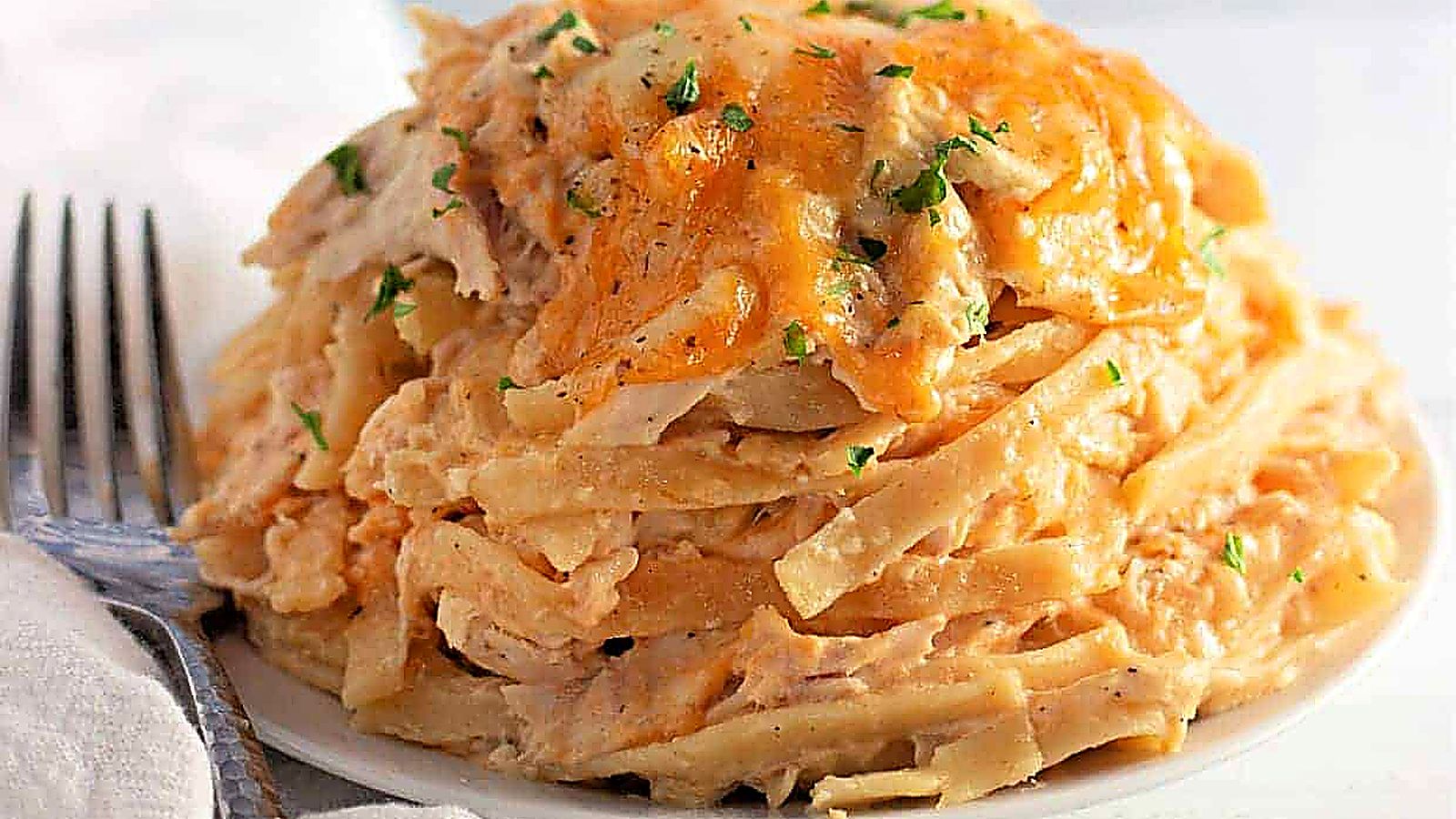 A plate of Chicken Tetrazzini topped with melted cheese and sprinkled with chopped herbs, with a fork and napkin beside it.