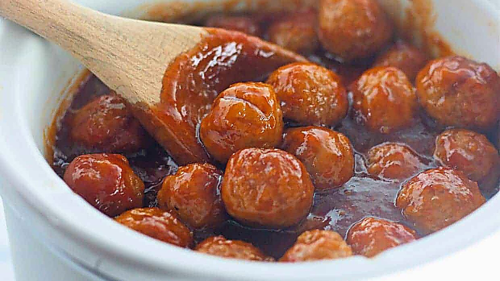 Honey-Garlic Meatballs in a thick red sauce are being stirred with a wooden spoon in a white ceramic pot.
