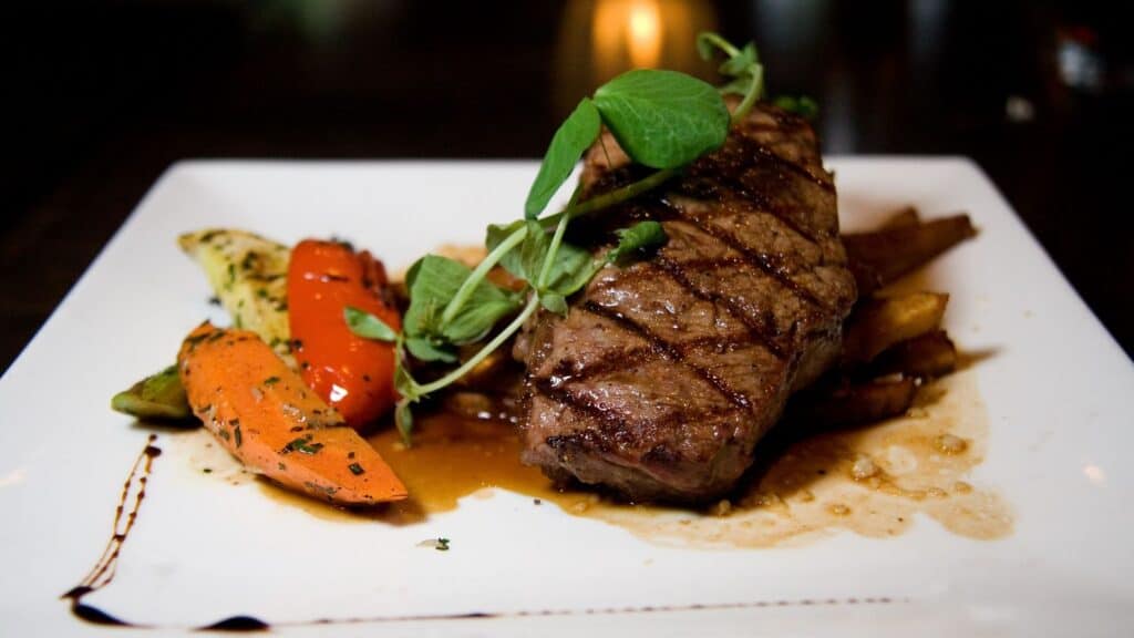 Grilled steak with grill marks served on a white plate alongside roasted vegetables and a rich brown sauce, garnished with fresh greens—a modern twist on mall food court staples elevated for a gourmet experience.