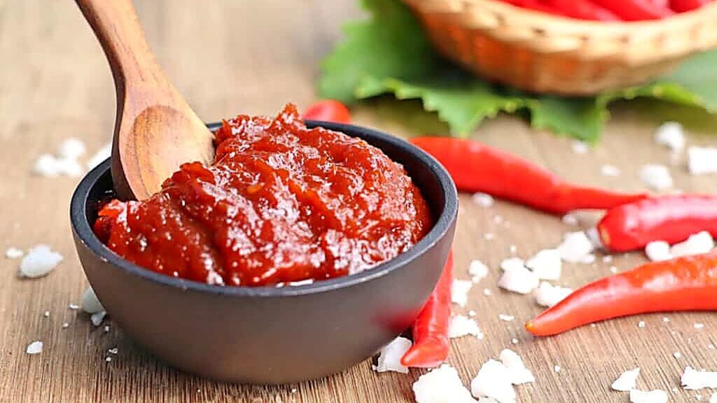 A black bowl of red chili paste with a wooden spoon, surrounded by red chili peppers and coarse salt on a wooden surface.