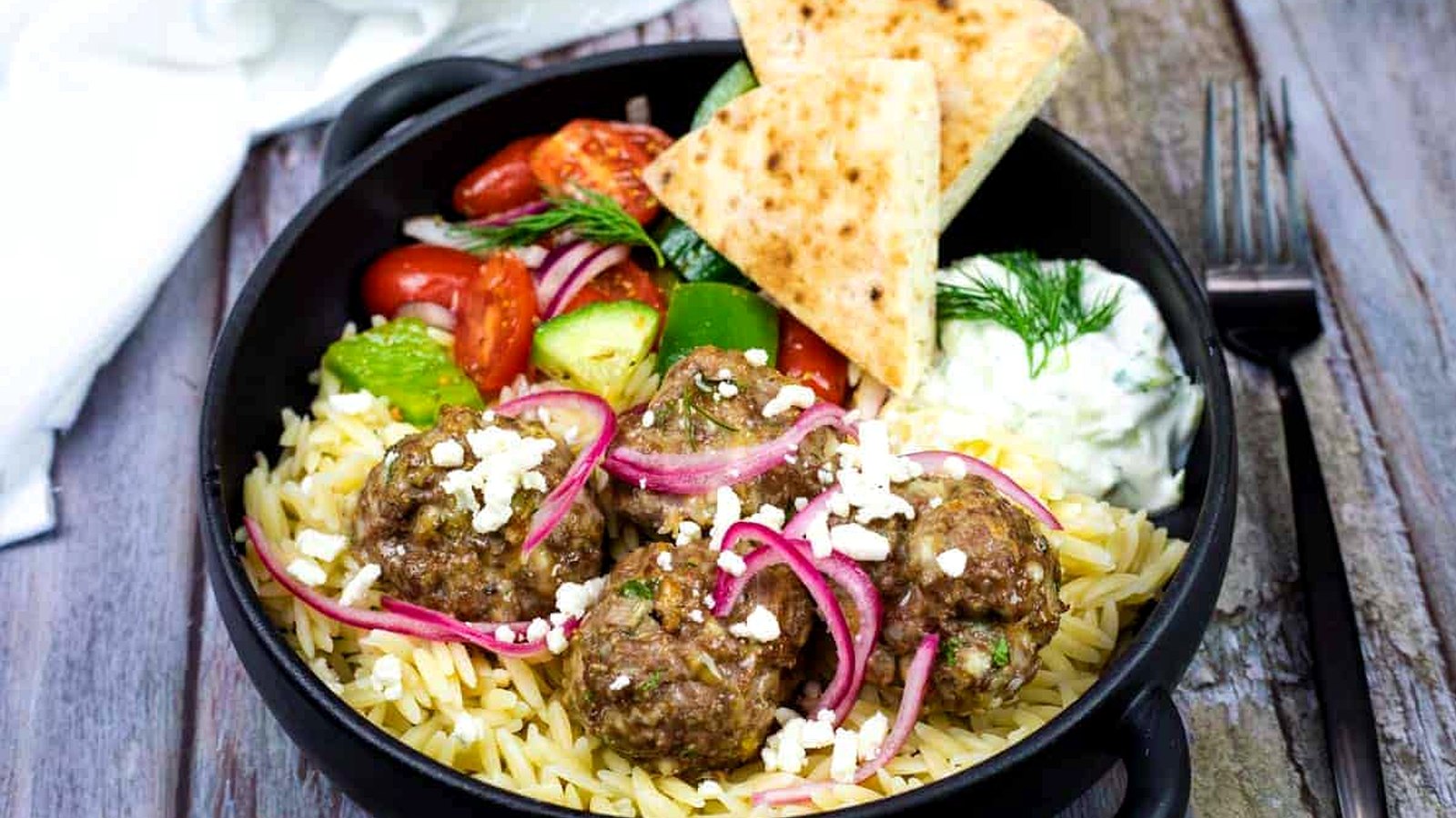 A black bowl with orzo pasta, meatballs topped with feta and pickled onions, Greek salad, pita bread, and a side of tzatziki sauce on a rustic wooden table.