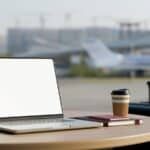 A laptop with a blank screen sits on a round table next to a coffee cup, notebooks, and a suitcase, creating the perfect setup to get work done in the airport, with an airport terminal and airplane visible through the window in the background.