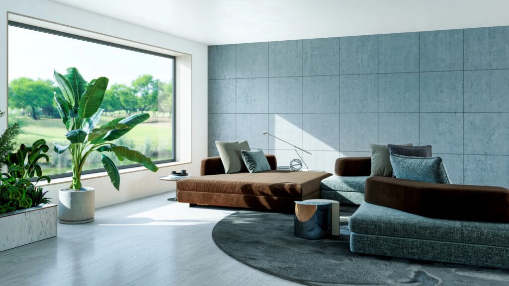 Modern living room with large window, brown and gray furniture, indoor plants, a round rug, and light wood flooring. Lush greenery is visible outside.