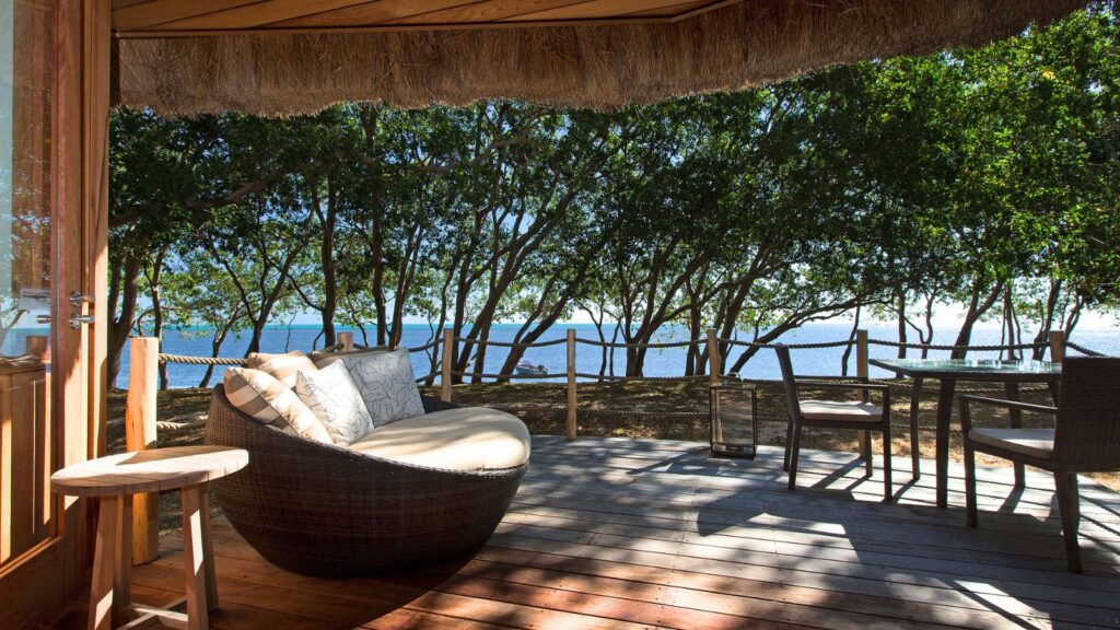 The wooden deck, part of the Marriott hotel collection, features a cushioned round chair and table overlooking trees and ocean through a rope fence, all bathed in clear daylight.