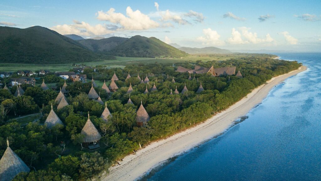 Aerial view of a tropical coastline with cone-shaped thatched huts among dense trees, mountains in the background, and calm blue ocean on the right.