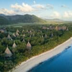 Aerial view of a tropical coastline with cone-shaped thatched huts among dense trees, mountains in the background, and calm blue ocean on the right.