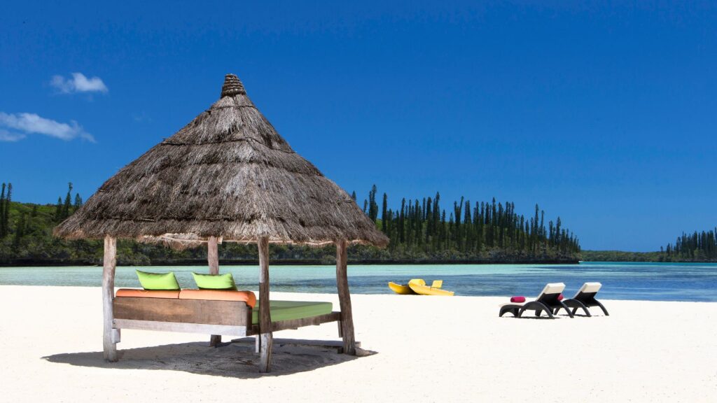A thatched-roof cabana with cushions, two lounge chairs, and two yellow kayaks sit on a sandy beach by calm blue water under a clear sky.