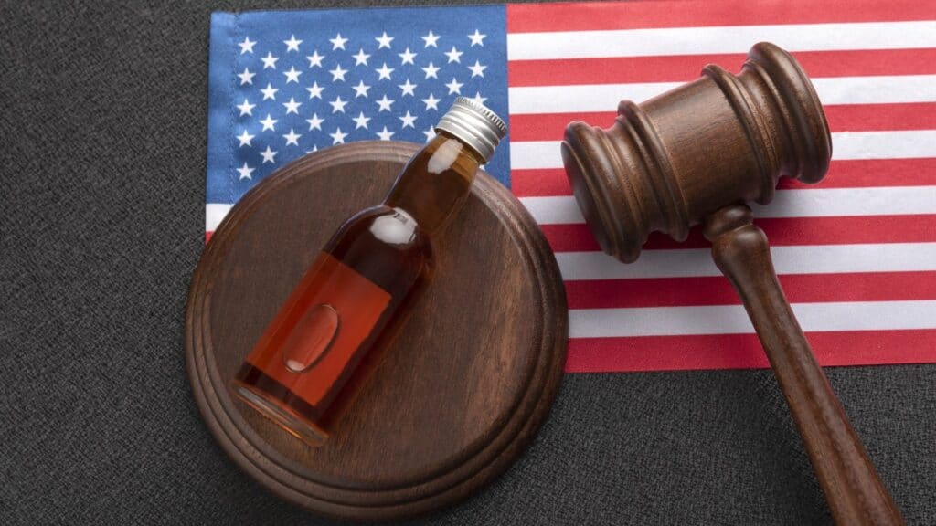 A small bottle of liquid and a judge&rsquo;s gavel rest on a wooden sound block, with a United States flag in the background, symbolizing the impact of state alcohol laws on legal proceedings.