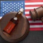 A small bottle of liquid and a judge’s gavel rest on a wooden sound block, with a United States flag in the background, symbolizing the impact of state alcohol laws on legal proceedings.